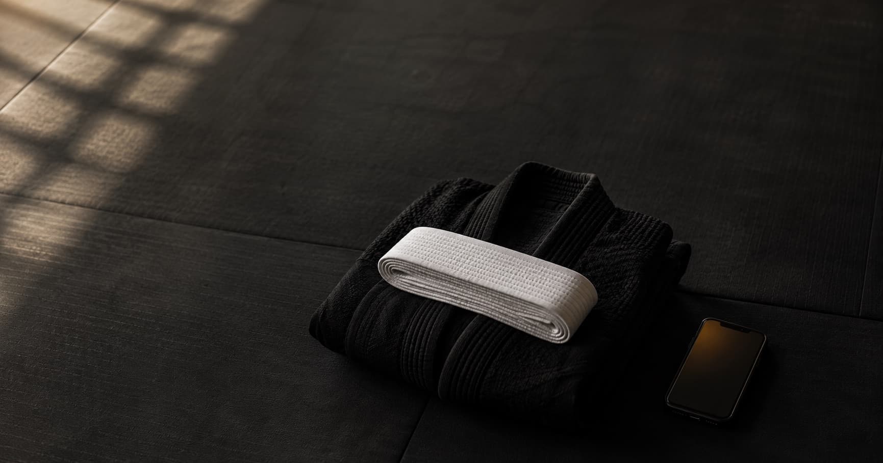 A folded white BJJ belt resting on a folded black gi top on a dark mat, with a phone laid flat beside them